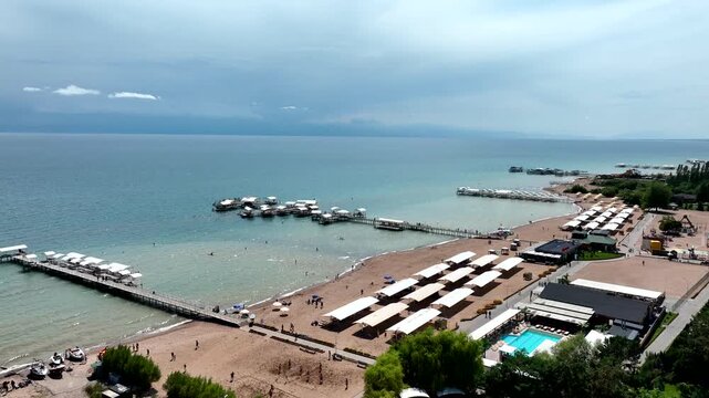 Aerial view of Issyk Kul lake with wooden piers, sandy beach, rows of white umbrellas, and resort facilities in Cholpon Ata, Issyk-Kul Region, Kyrgyzstan.