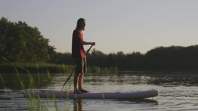 Sup board boad man. A man on a paddleboard navigates the calm water while surrounded by greenery. The man maintains balance on the paddleboard amidst the serene landscape.