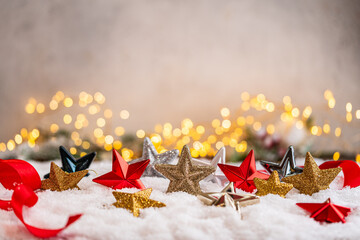 Naklejka premium A Christmas background featuring an array of red, gold, silver, and navy glittering stars scattered on snow beside a red ribbon, softly lit by warm twinkling lights with copy space.