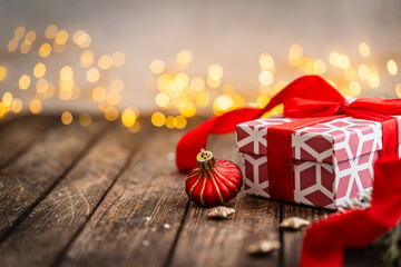 Naklejka premium A Christmas background featuring a red and white geometric-patterned gift box tied with a satin bow beside a striped ornament and golden accents on rustic wood, lit by warm twinkling lights.
