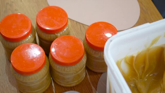 Cinematic close-up tracking shot moving from right to left from retail honey jars with orange lids to a bulk bucket of thick raw mountain honey.