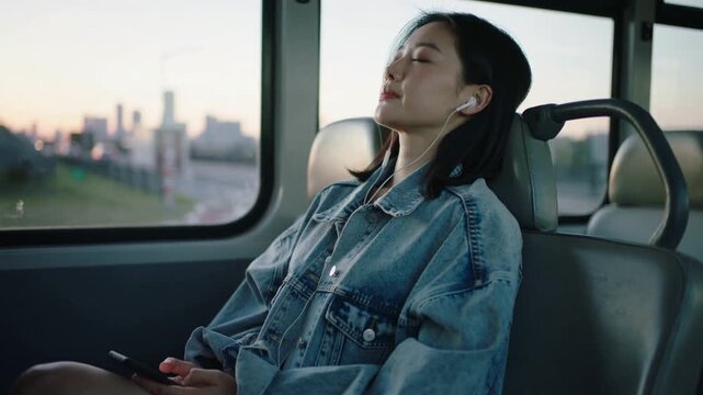 Relaxing woman wearing denim jacket holding smartphone, wired earphones as bus lulling past skyline
