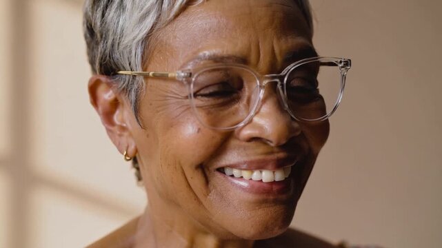 Smiling senior woman at home responding to light widening smile, clear frames, gold hoops, window