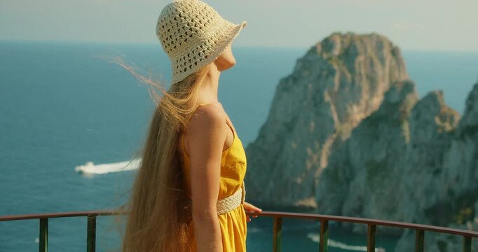 Woman enjoying windy cliffside sea view of Faraglioni rock formations in summer Capri