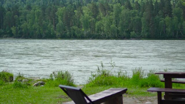 Cinematic smooth push-in footage moving forward past an empty riverside campfire picnic zone with a wooden table, benches, and a grill towards the fast-flowing Katun River in the Altai Mountains, Sibe