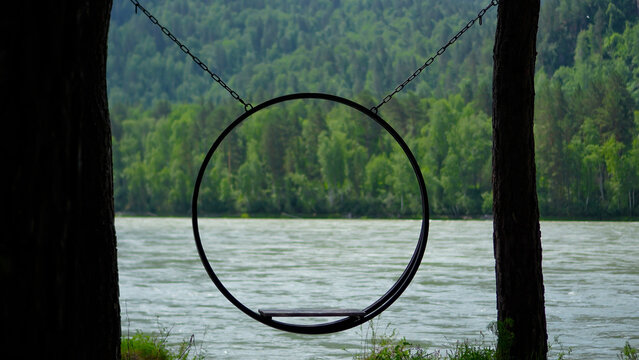 Artistic silhouette frame of a modern circular hanging swing on chains with a beautiful background of the fast-flowing Katun River and dense Siberian pine forests.