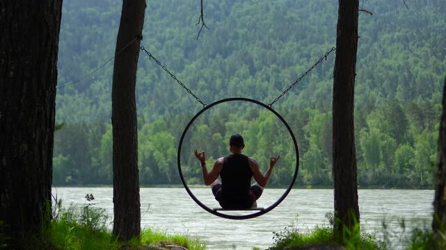Inspiring static footage from a back view of a male traveler sitting in a lotus pose on a circular hanging swing, meditating over the Katun River in the Altai Mountains, Siberia.