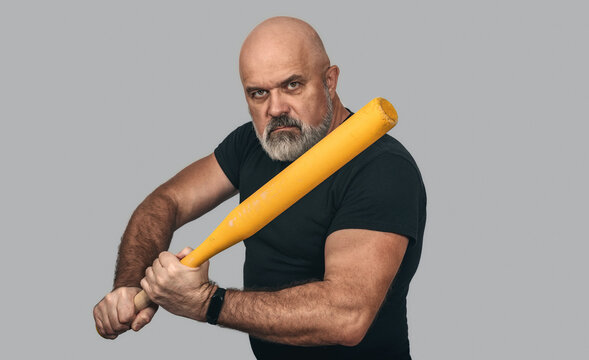 Bearded strong man holding baseball bat in intimidating pose. Gray background, isolated. Aggression.
