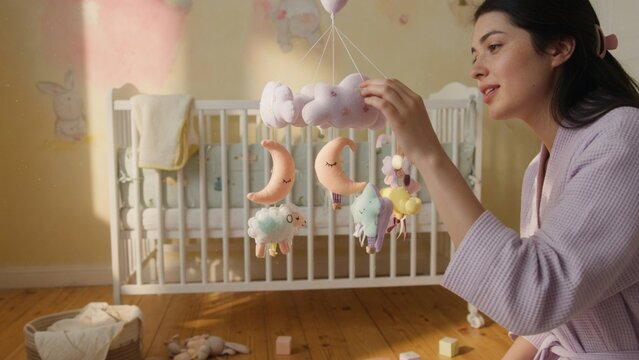 Young mother in cozy pastel nursery adjusting soft baby crib mobile with plush clouds, moons, and sheep toys bathed in warm golden sunlight at tender parenting moment