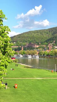 People walk along the riverbank while boats pass by on a sunny day in a green park
