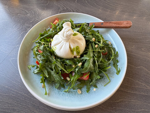 Creamy Burrata Rests On Peppery Arugula With Tomato Pieces, Toasted Nuts, And Green Dressing In Shallow Bowl On Dark Wooden Surface, Presenting Fresh Italian Meal For Restaurant Menu Or