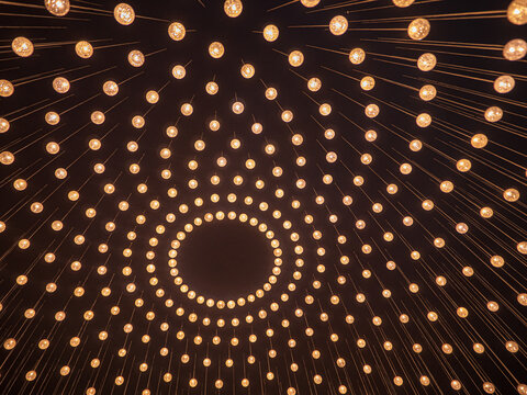 Many warm glowing lights arranged in concentric circles, creating a symmetrical radial pattern against a dark background. Abstract geometric composition with golden illumination.