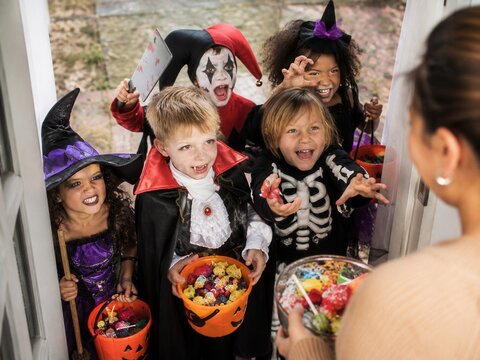 Kindergruppe sammelt S&uuml;&szlig;igkeiten an der Haust&uuml;r. Verkleidete Kinder in Halloween Kost&uuml;men halten Eimer voller Bonbons, w&auml;hrend eine Frau an der T&uuml;r S&uuml;&szlig;es verteilt.