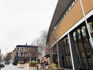 Naklejka premium Modern city street with contemporary office building and historic pub facade, pedestrians and planters in London, UK on an overcast winter day
