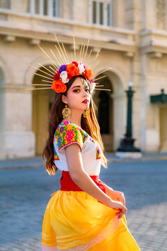 Young latina woman portraying a Catrina with makeup and a floral halo crown, wearing a vibrant embroidered blouse and yellow skirt while posing in front of a historic building in Havana