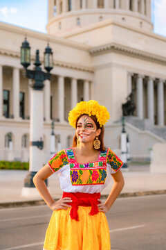 Young Latina woman with traditional Catrina skull makeup and a flower crown, wearing an embroidered dress, celebrating Day of the Dead in front of the Capitolio building in Havana, Cuba
