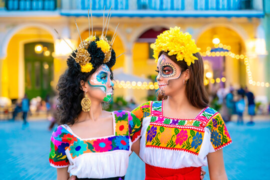 Two women wearing vibrant catrina makeup and traditional embroidered dresses celebrating the Day of the Dead tradition, featuring colorful face paint, floral headpieces, and festive attire