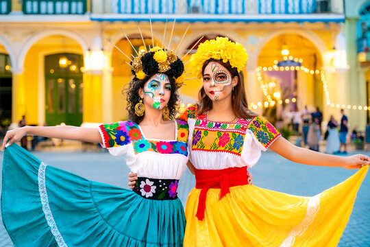 Two young Latin women wearing traditional Mexican blouses and vibrant flower crowns, with Catrina skull makeup on their faces, posing outdoors during a cultural festival