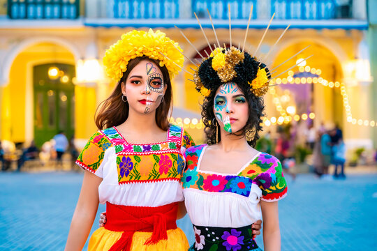 Two young women, dressed in traditional embroidered blouses and colorful flower crowns, celebrating the Day of the Dead with half face skull makeup, standing on a street in Havana, Cuba