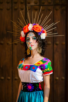 Latina woman with traditional Catrina makeup and a vibrant floral crown, wearing an embroidered Mexican dress, posing against a wooden door, celebrating Mexican traditions in Cuba