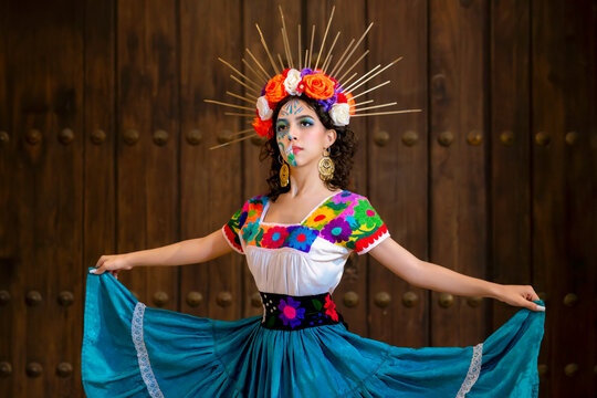 Latina woman celebrating Day of the Dead, wearing vibrant embroidered folk costume, skull makeup, and a floral crown with golden rays, posing against a wooden door