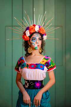 Latina woman with intricate sugar skull makeup, wearing a colorful embroidered dress and floral headdress with a halo, honoring Mexican traditions for Dia de Muertos