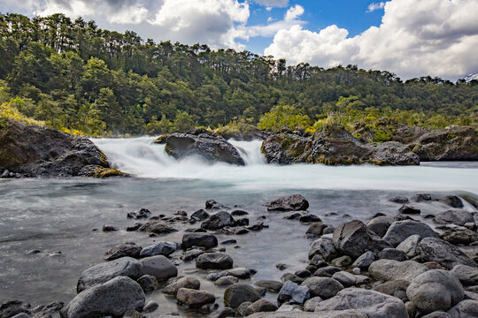 Scenic cascading waterfall over dark volcanic rocks with surrounding lush forest and cloudy blue sky