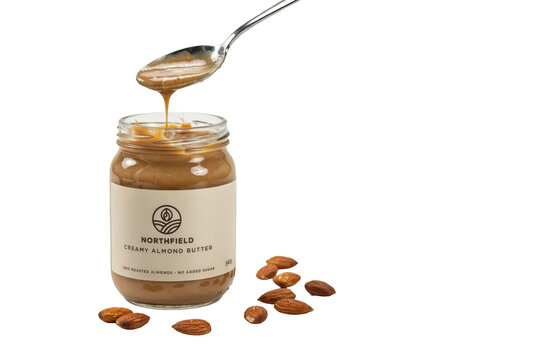 Glass jar of creamy almond butter with a silver spoon dripping nut butter and scattered whole almonds on white isolated on a transparent background
