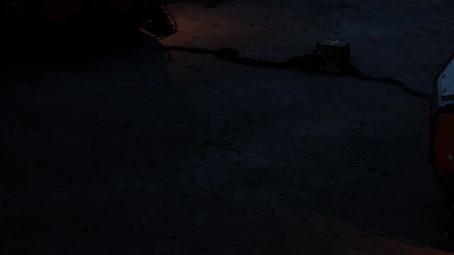 A quiet street scene shows dim street lights shining on the pavement during the evening. Shadows extend from objects on the ground. The setting appears urban and somewhat deserted.