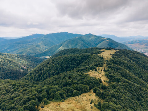 Sweeping drone panorama of rolling forest-covered ridges, open mountain meadows and distant valleys under an overcast sky.