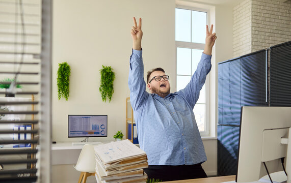 Overweight man dancing in office by computer. He raises arms in victory near charts, celebrating business progress and better financial results. Clear concept of office success and joy.