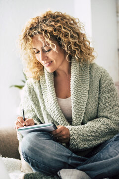 A relaxed woman in a cozy sweater writing in a notebook at home, smiling thoughtfully as she journals on a comfortable couch