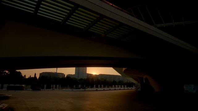 Sunset urban plaza beneath sweeping curved overpass, dark architectural spans above open paving, distant high-rise tower, rows of trees, warm amber sky, deep shadow, modern geometry and calm city