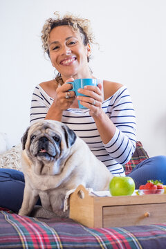 Smiling woman enjoys a morning coffee at home with her pug sitting on a cozy bed beside a breakfast tray with an apple and strawberries, conveying relaxation, companionship and simple domestic joy.