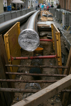 Wrapped pipeline in city trench, foil insulation and protective cap over metal pipe, muddy base, adjacent conduit lines and cable bundles. Urban construction site, municipal maintenance