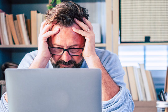 Middle-aged man wearing glasses rests his head in his hands while looking at a laptop in a home office, conveying stress, frustration and fatigue from remote work or long hours