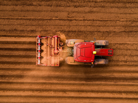Red tractor pulling a planter, creating rows while seeding carrots in rich brown soil. Aerial view of modern farming
