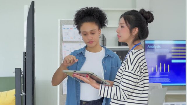 Two focused female colleagues reviewing information together on a tablet, one explaining a point while wearing a lanyard, in a bright collaborative office space.