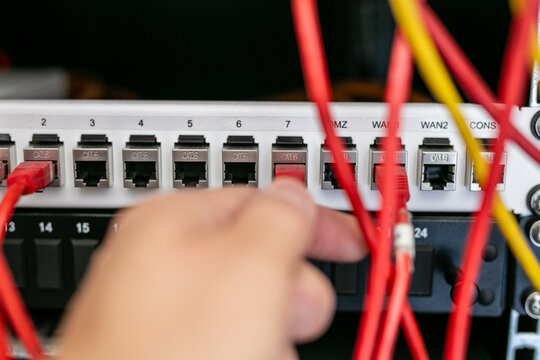 Close-up of a person's hand plugging an Ethernet patch cable into a numbered network port on a patch panel or network switch inside a server rack. The image illustrates structured network cabling