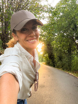 Smiling woman taking a sunset selfie on a quiet country road, casual cap and shirt, sunglasses hanging, enjoying nature and a peaceful evening walk