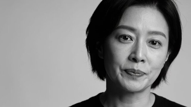 Turning Asian woman speaking to camera in studio, giving speech, plain backdrop, wearing black top