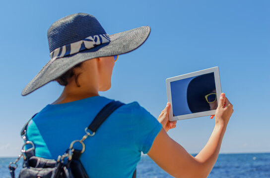 A woman in a sun hat and blue shirt uses a digital tablet near the ocean on a bright sunny day.
