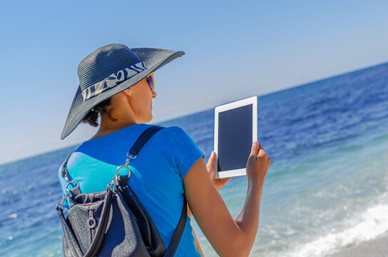 A woman in a sun hat holds a digital tablet while standing on a sunny beach looking out at the ocean.