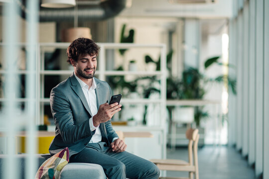 Young professional businessman smiling, using smartphone for communication and networking in modern office lounge area