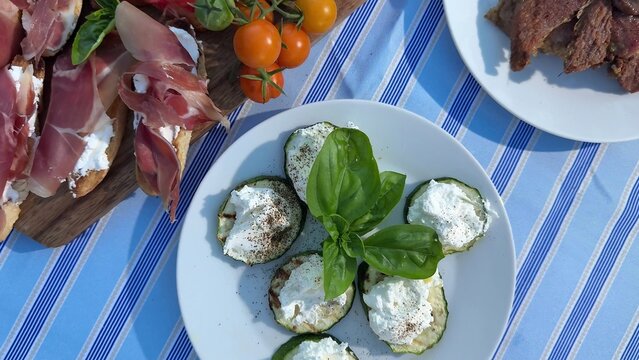 Grilled zucchini slices topped with fresh ricotta cheese and basil, Mediterranean appetizer platter on blue striped tablecloth, bright summer daylight
