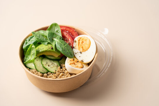 Healthy protein bowl with egg, avocado, spinach, cucumber and quinoa in a cardboard container on a light beige background, nutritious balanced meal concept for healthy eating