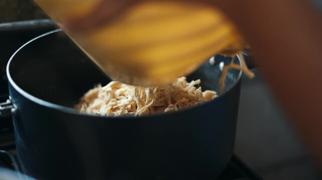 Close up of shredded chicken meat being poured from a bowl into a pot for cooking a family meal tinga