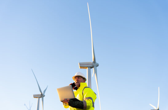 Engineer talking on phone and holding laptop, working at a wind farm, managing renewable energy production