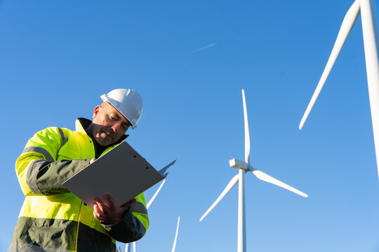 Engineer wearing a hard hat and high-visibility jacket, writing on a clipboard while standing among wind turbines