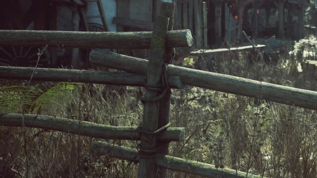 A wooden fence made of logs stands in a rural area. The grass is tall and unkempt around the fence. A structure is visible in the background. The sun shines on the scene, creating shadows.